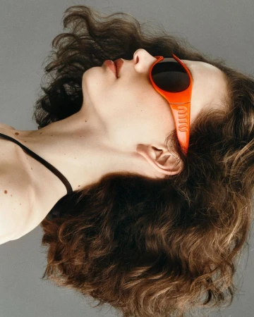 Model Sean Levy in profile with head tilted back, wearing bright orange wraparound sunglasses against a grey studio backdrop.