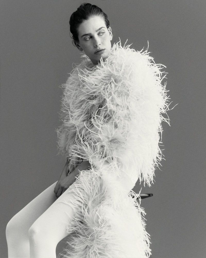 Model Sean Levy seated wearing an oversized white feather coat and white tights against a plain studio background.