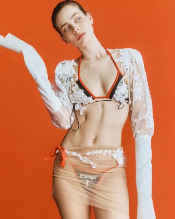 Model Sean Levy posing in a white lace shrug, bikini top, and sheer skirt against a bold solid orange background.