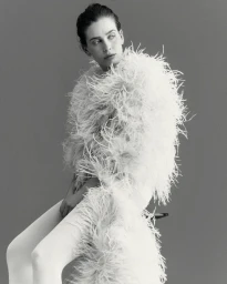Model Sean Levy seated wearing an oversized white feather coat and white tights against a plain studio background.