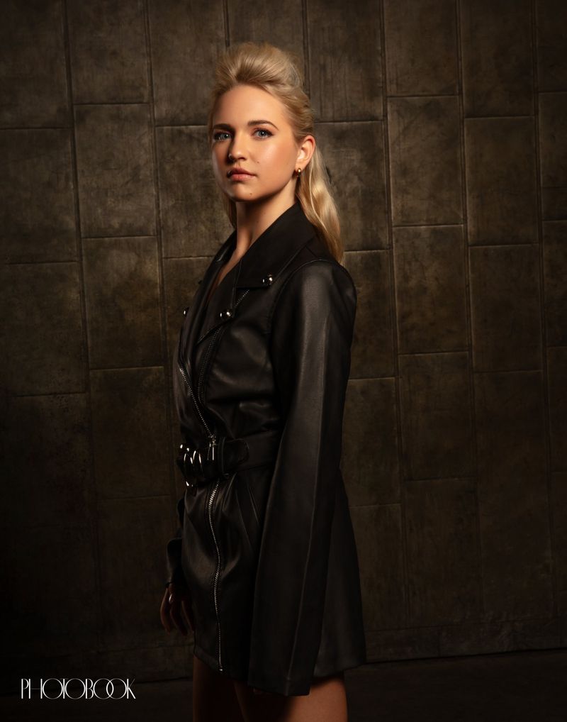 Blonde woman wearing a black leather coat against a dark tiled stone wall in a portrait for Photobook Magazine