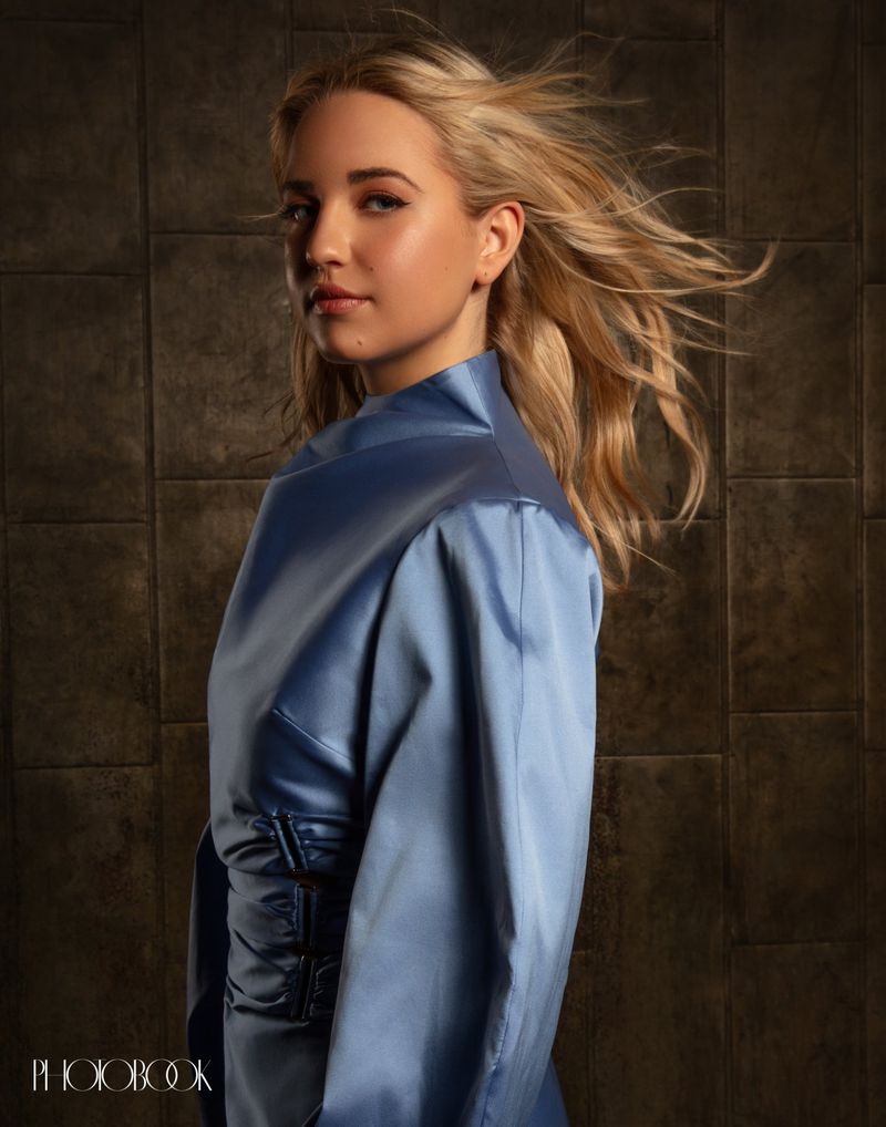 Portrait of a blonde woman wearing a blue satin high-neck top against a dark tiled background for Photobook Magazine.