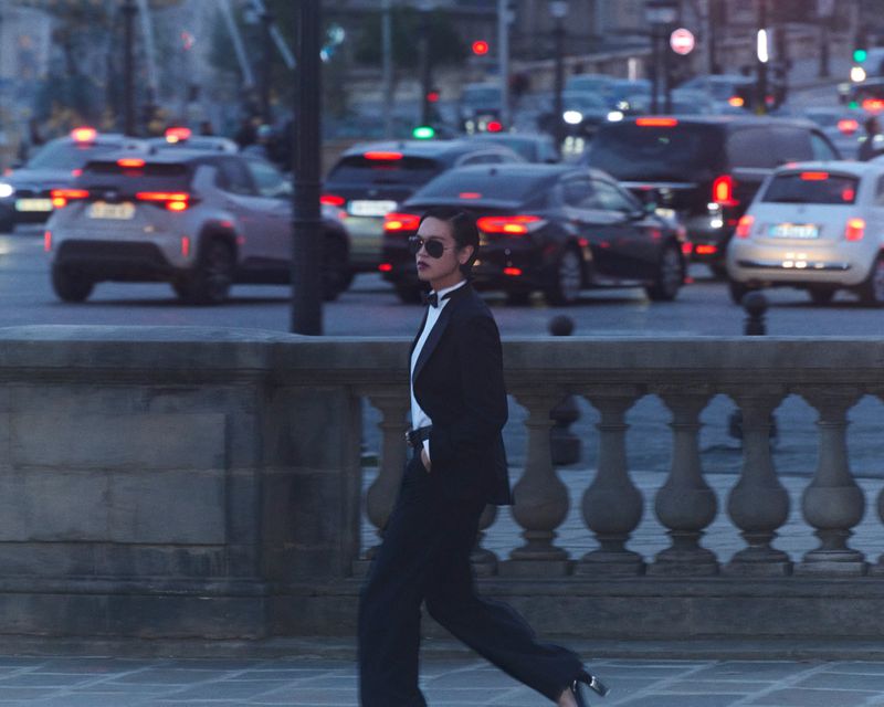 Model Dara walking across a stone bridge wearing a black tuxedo suit, bow tie, and sunglasses with city traffic behind.