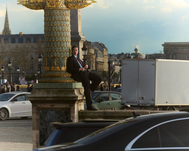 Model Dara wearing a black suit and leaning against an ornate stone column in a Parisian public square with passing city traffic.