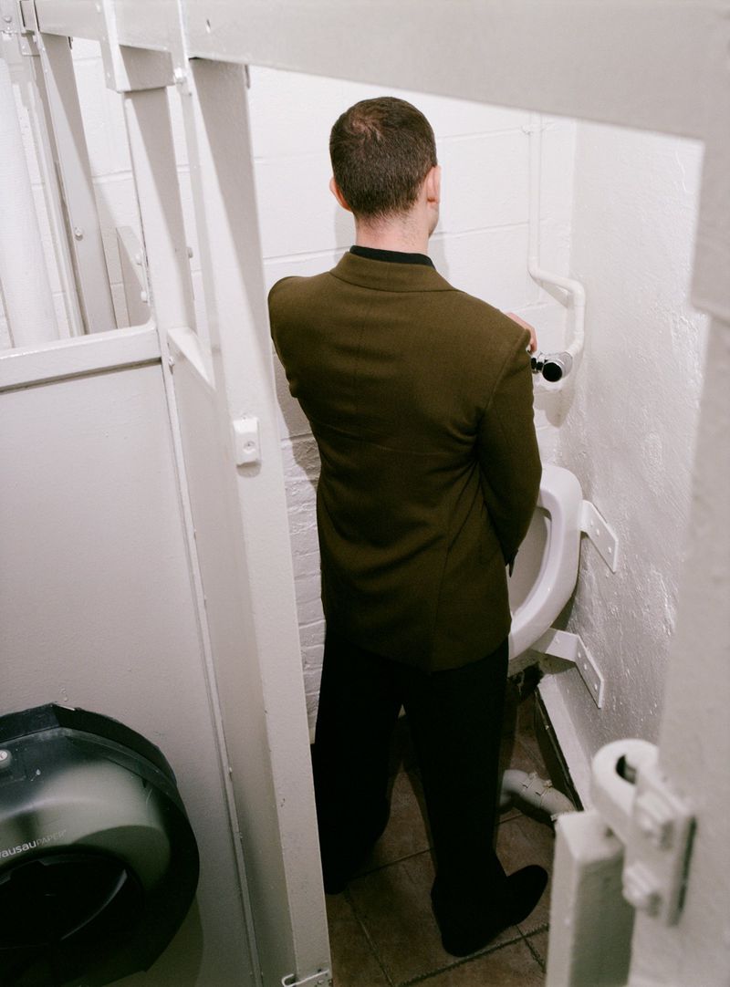 Back view of a man in an olive green blazer and black trousers standing at a white urinal inside a public restroom stall.