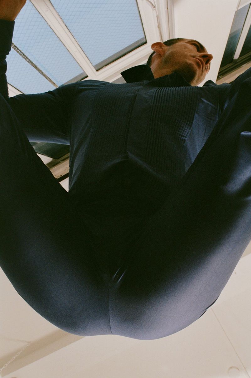 Low-angle shot looking directly up at a man wearing a dark navy pleated dress shirt and trousers positioned below a window.