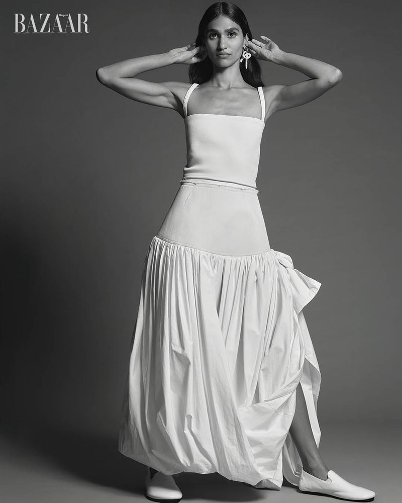 Hannah Wick poses in a white square-neck top, drop-waist gathered maxi skirt, and white flats against a studio backdrop.