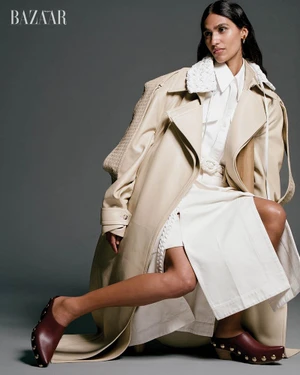 Model Hannah Wick poses in an oversized beige leather trench coat and white skirt against a studio backdrop.