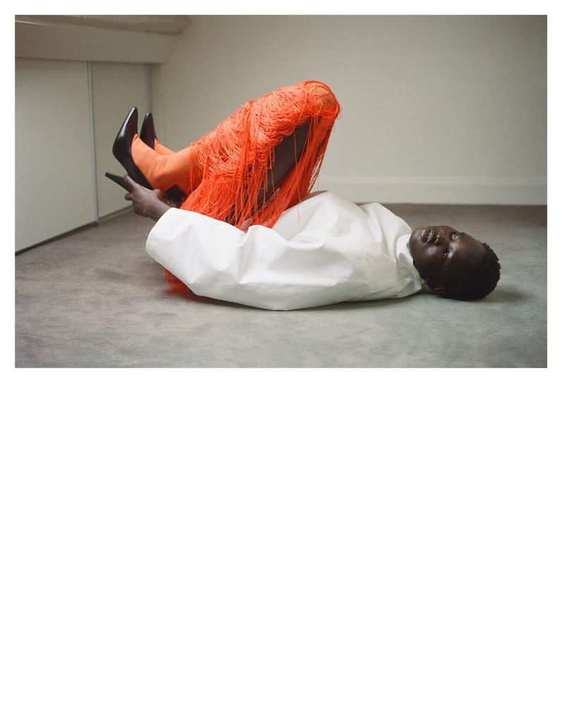 Model Nyakong Chan lies on a grey carpet with legs raised, wearing an oversized white top, vibrant orange fringe pants, and black heels.