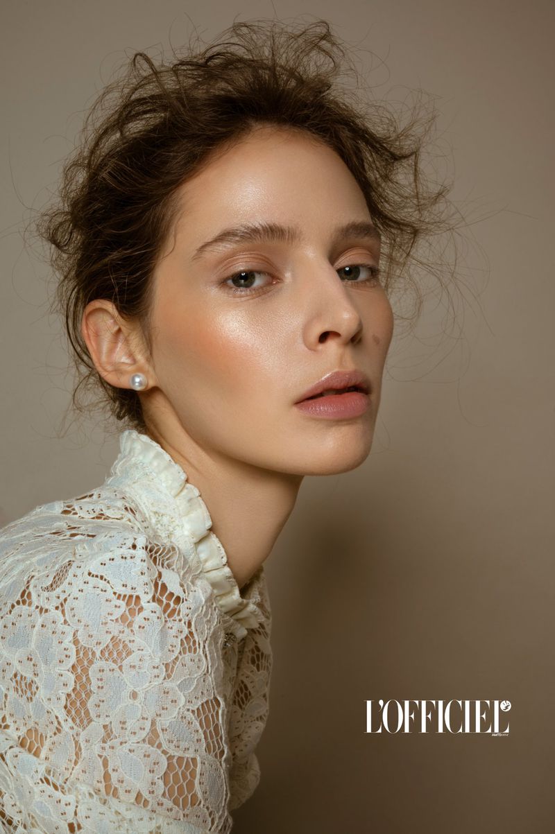 Model Alisa Körbitz in a high-neck white lace top and pearl earring against a neutral beige background for L'Officiel Belgium.