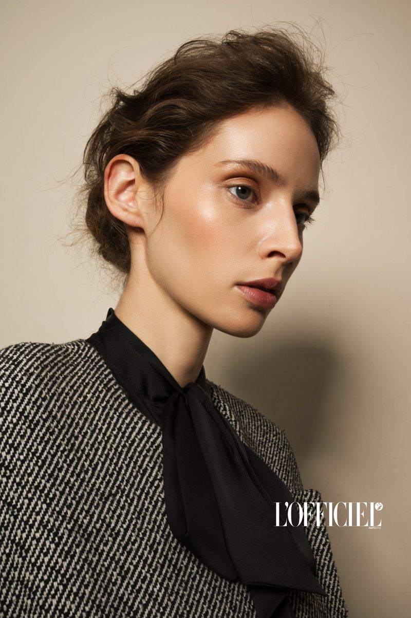 Model Alisa Körbitz in profile wearing a tweed blazer with a black bow blouse against a warm neutral background.