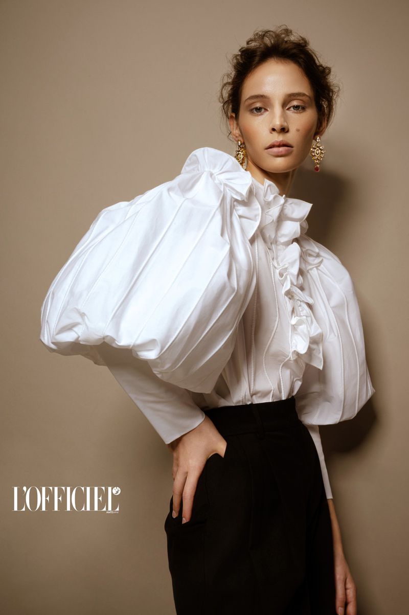Model Alisa Körbitz wearing an exaggerated white puff-sleeve ruffle blouse and black trousers against a beige backdrop.