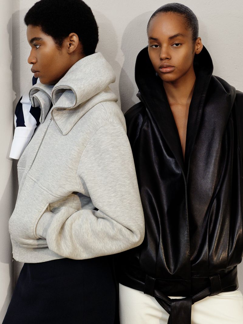 Two models standing side-by-side wearing sculptural outerwear including a grey fleece jacket and a black draped leather coat.