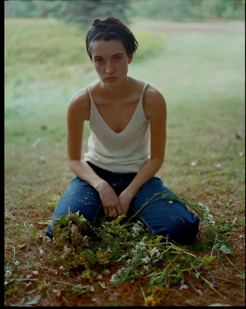 Kit Pugh kneeling outdoors in a white tank top and blue jeans behind scattered wildflowers for Polène Paris.