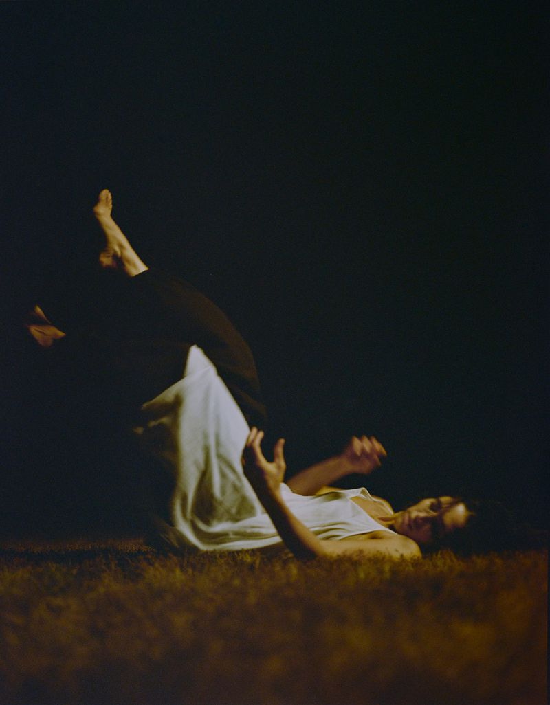 Model lying on grass at night with legs raised wearing a white top and dark pants under dramatic low-key lighting.