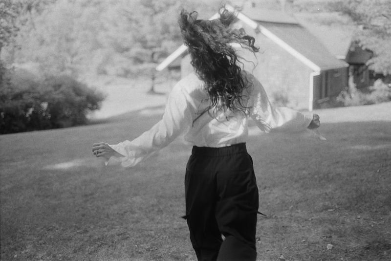 A person with long dark wavy hair seen from behind running across a lawn wearing a white blouse and black trousers.