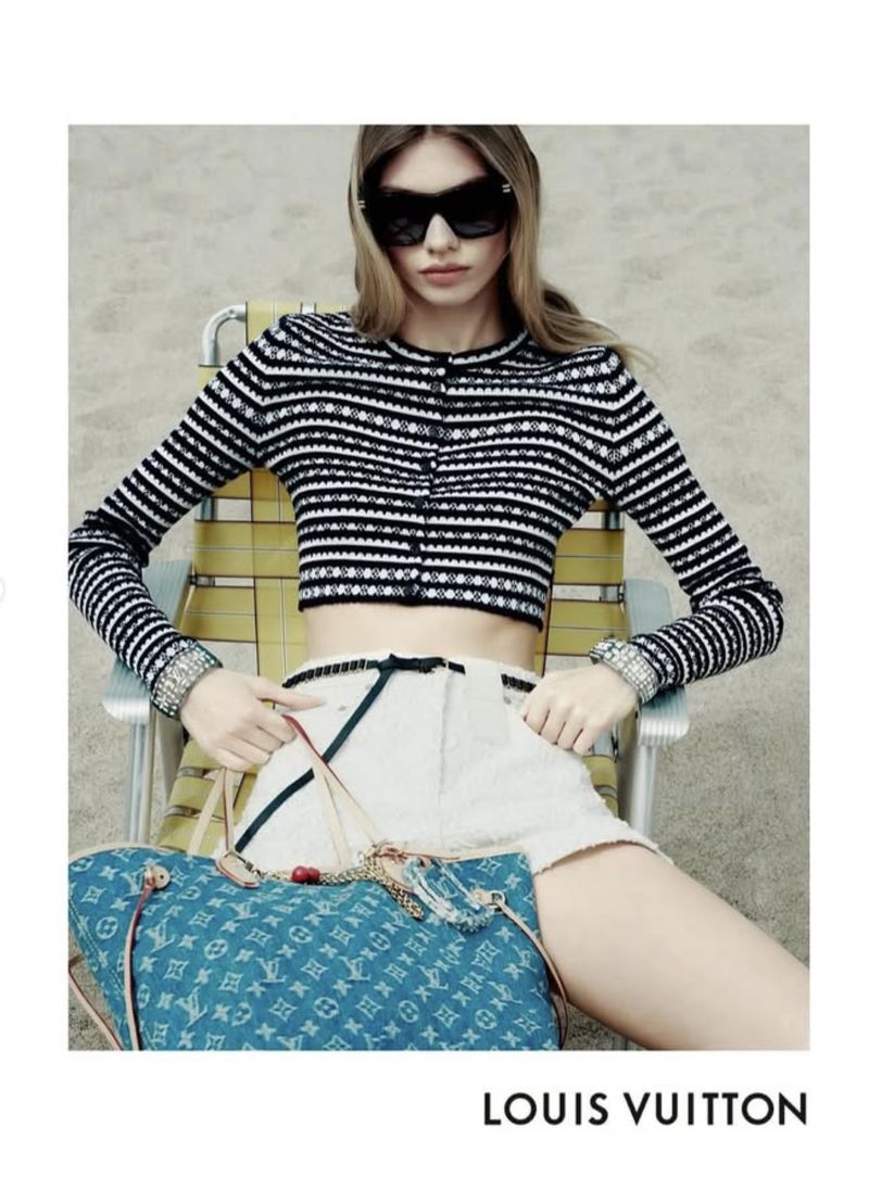 A fashionable adult female in a striped sweater, sunglasses, and shorts is sitting outside with a blue Louis Vuitton purse.