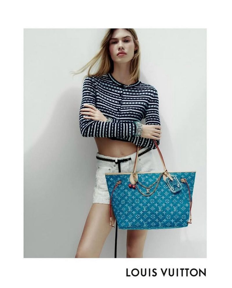 A blonde fashion model poses in a cropped striped shirt and white shorts, holding a Louis Vuitton handbag against a white wall.