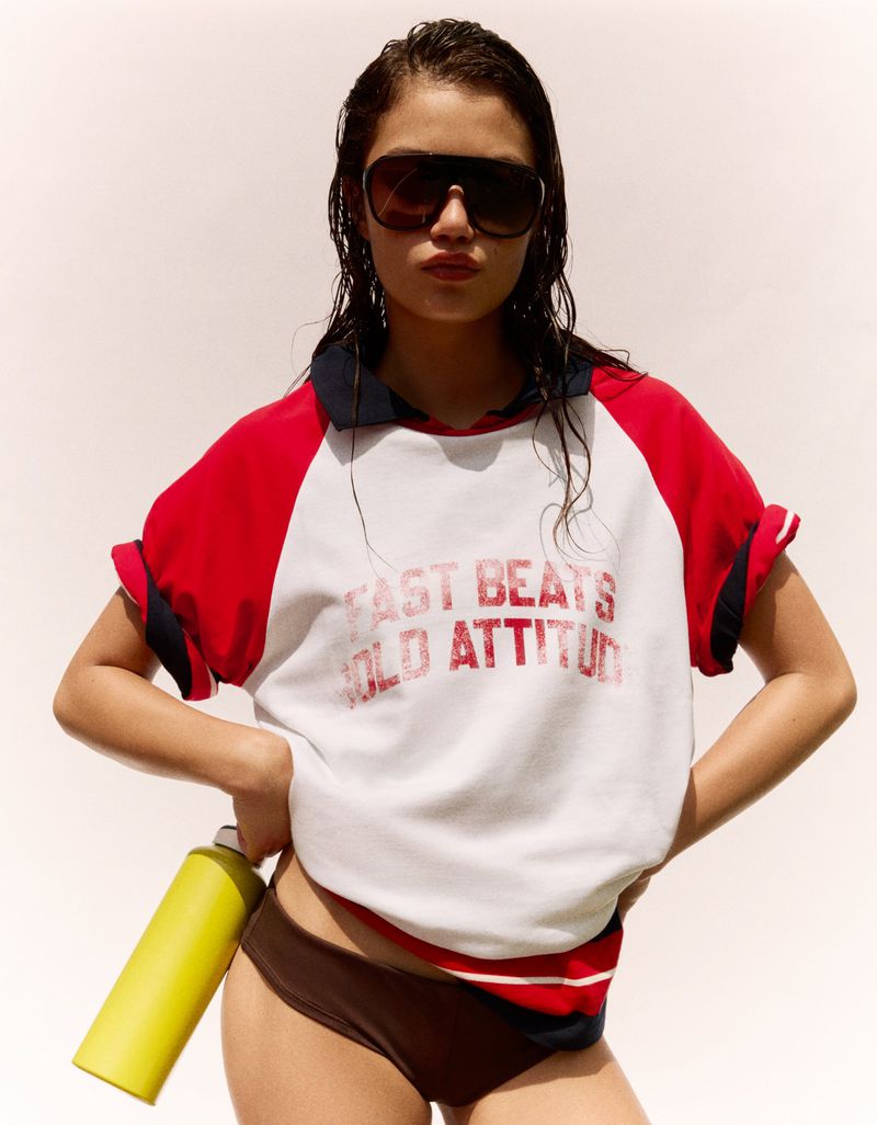 Model wearing oversized shield sunglasses, a red and white graphic polo top, and holding a yellow bottle against a white backdrop.