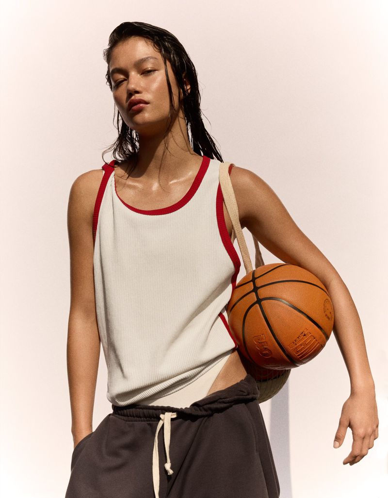 Model wearing a white ribbed tank top with red trim and dark sweatpants holding a basketball against a neutral background.