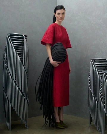 Model in red midi dress holding a black sculptural accessory between two stacks of metal chairs against a textured grey wall.
