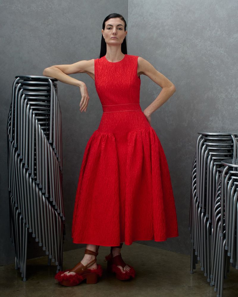 Model Eliana Weirich in a bright red sleeveless midi dress leaning against stacked metal stools in a grey studio corner.