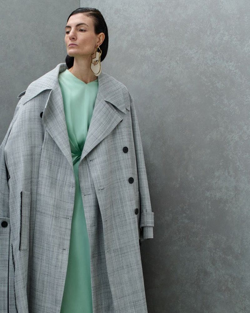 Model Eliana Weirich wearing an oversized grey plaid trench coat and mint green dress against a textured grey wall.