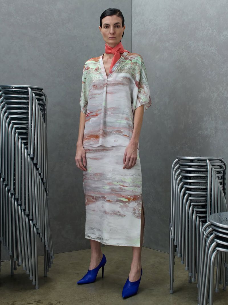 Eliana Weirich wearing a printed two-piece midi set, coral neck scarf, and cobalt blue heels beside stacks of metal stools.
