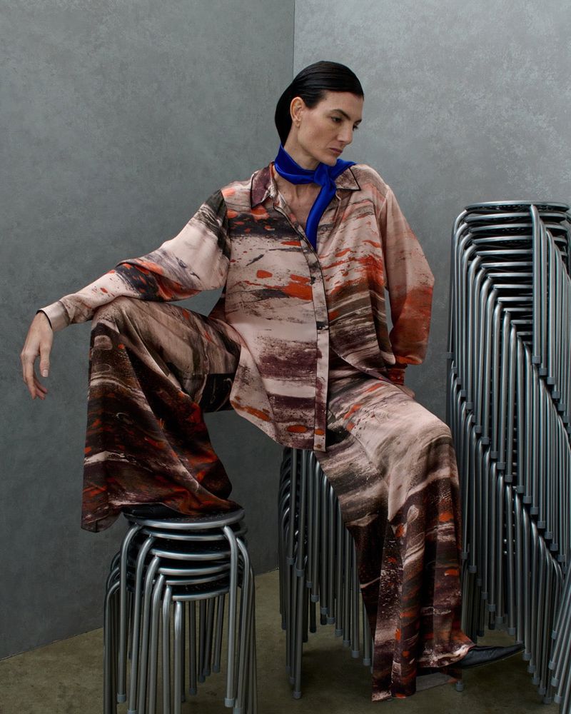 Model Eliana Weirich seated on a stack of metal stools wearing a printed silk shirt and wide trousers with a blue neck scarf.