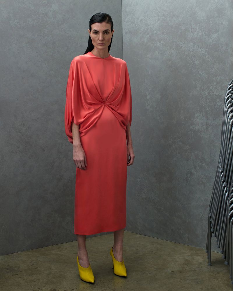 Model Eliana Weirich wearing a coral draped midi dress and yellow pointed heels against a grey textured studio backdrop.