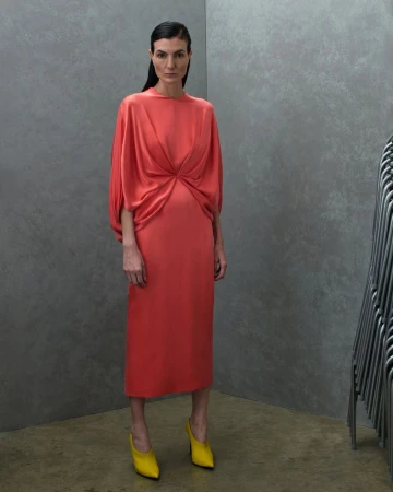 Model Eliana Weirich wearing a coral draped midi dress and yellow pointed heels against a grey textured studio backdrop.