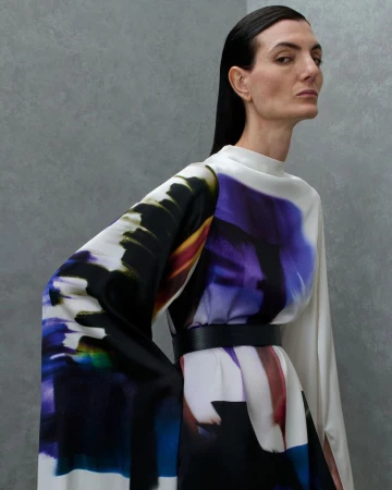 Model Eliana Weirich wearing a high-neck draped dress with abstract purple, blue, and black prints belted at the waist against grey.
