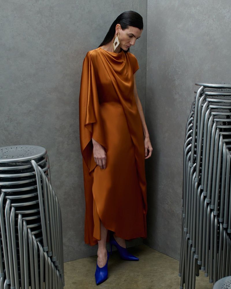 Model Eliana Weirich in a draped copper satin gown and cobalt blue heels standing between stacks of metal chairs.