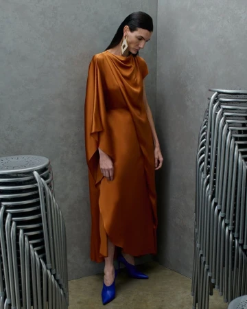 Model Eliana Weirich in a draped copper satin gown and cobalt blue heels standing between stacks of metal chairs.
