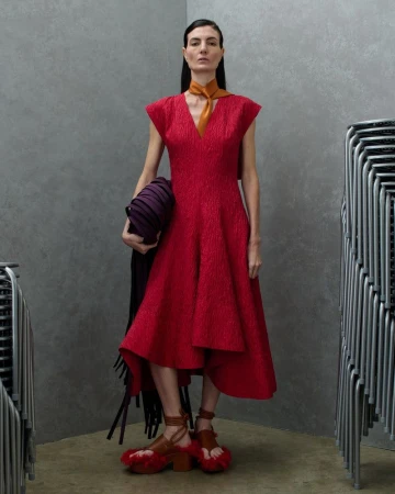 Model wearing a sleeveless crimson red midi dress and mustard neck scarf standing between stacks of metal chairs in a studio corner.