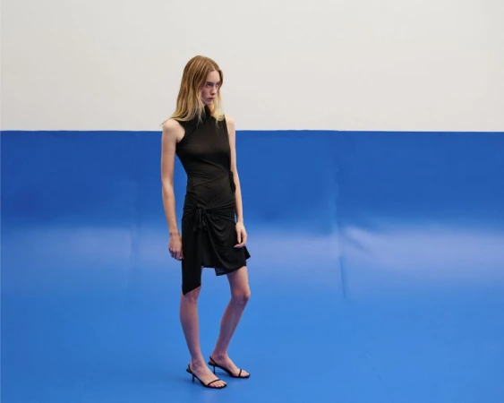 Model standing against a two-tone blue and white backdrop wearing a black sleeveless dress and sandals.