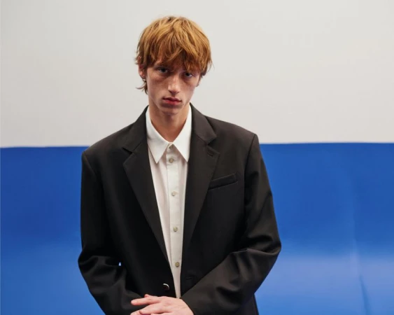 Model Oscar Fishman in a black blazer and white collared shirt against a two-tone blue and white studio backdrop for Helmut Lang.