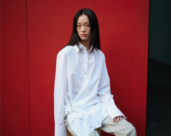 Model Jiayi Liang seated wearing an oversized white draped button-up shirt and light pants against a vibrant red backdrop.