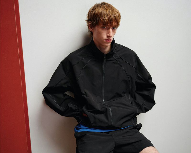 Male model Oscar Fishman wearing a black contrast-stitch zip-up jacket and matching shorts leaning against a white studio wall.