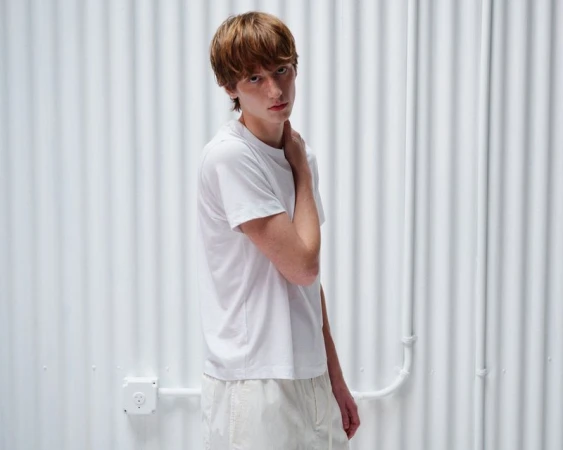 Young male model wearing a white t-shirt standing against a white corrugated metal wall for Helmut Lang Pre-fall 2026
