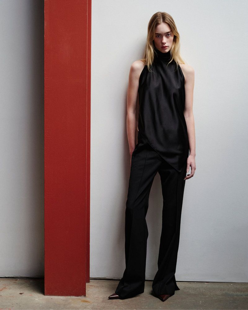 Model wearing a black sleeveless draped halter top and matching tailored black trousers standing beside a red column.