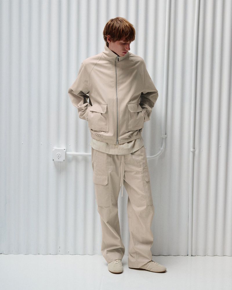 Model wearing a beige zip-up jacket and matching cargo trousers standing against a white corrugated wall.