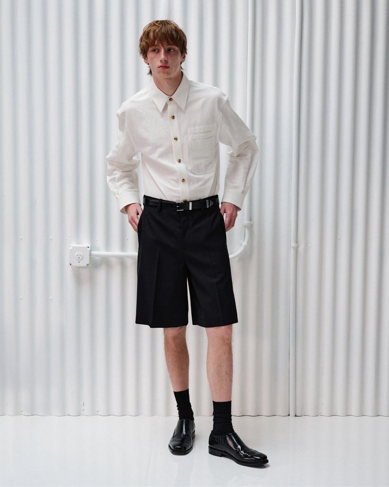 Oscar Fishman in a white button-up shirt and black pleated shorts against a white corrugated backdrop for Helmut Lang.