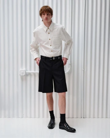Oscar Fishman in a white button-up shirt and black pleated shorts against a white corrugated backdrop for Helmut Lang.