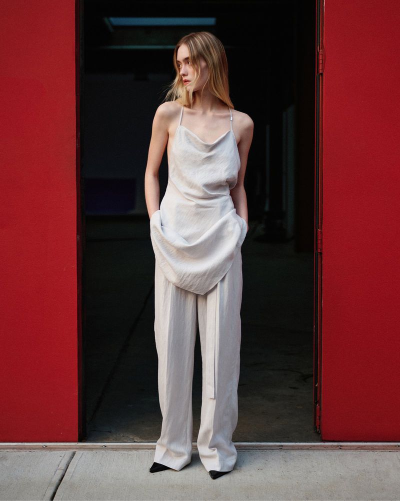 Model standing in a red doorway wearing a cream draped cowl neck top and wide leg matching trousers.