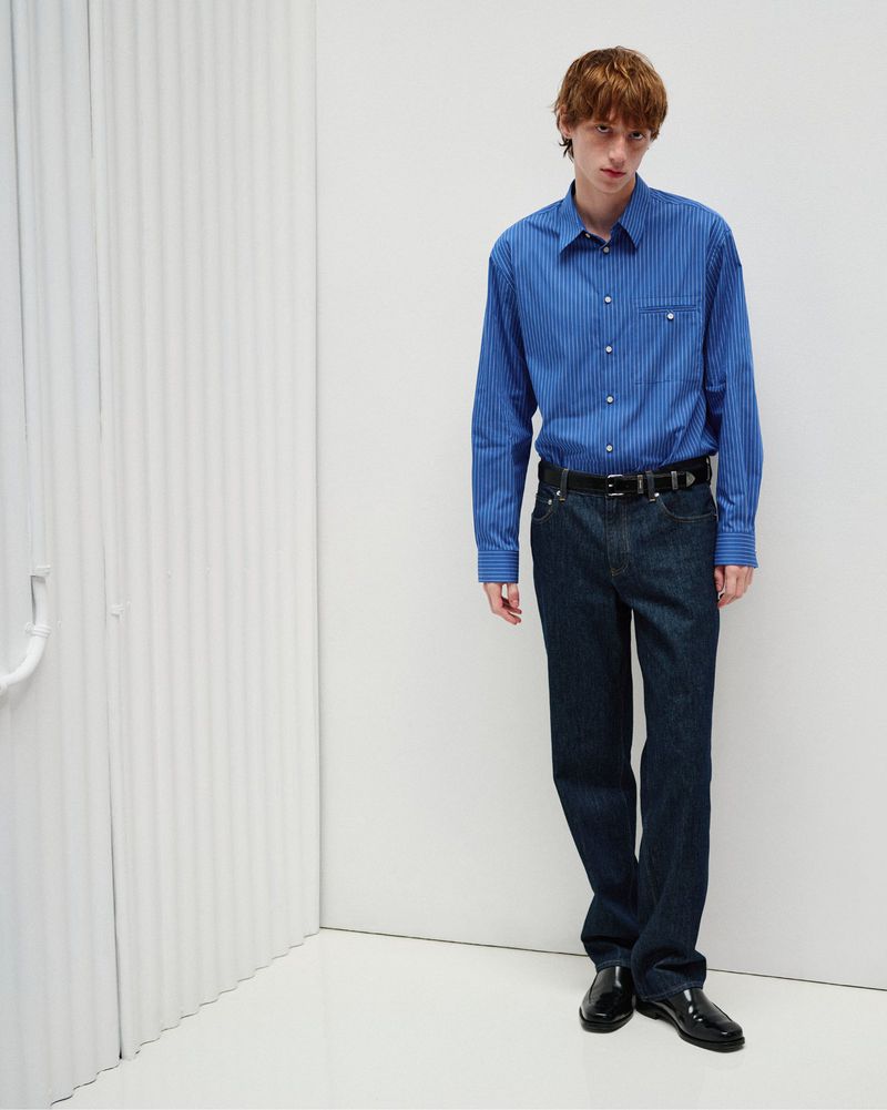Model standing in a blue button-down shirt and dark denim jeans against a white ribbed studio backdrop.