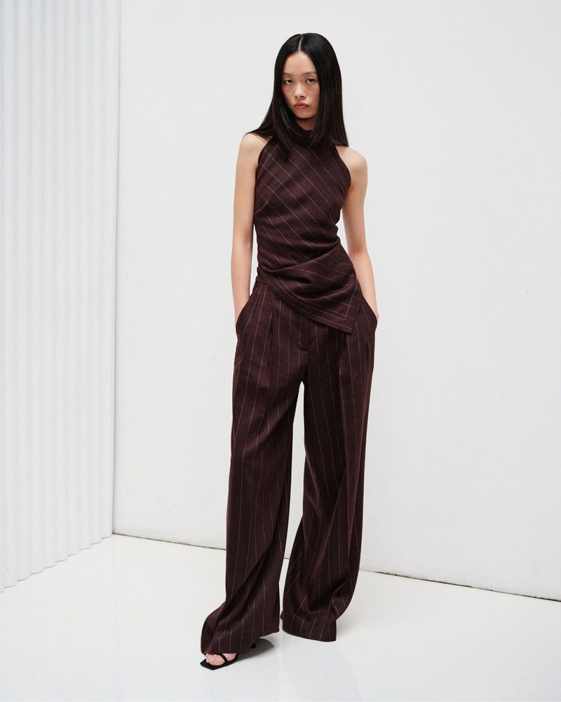 Model wearing a dark brown pinstriped sleeveless draped top and matching wide-leg trousers in a white minimalist studio.