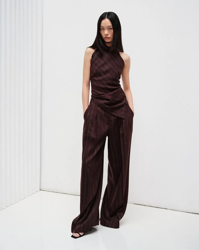 Model wearing a dark brown pinstriped sleeveless draped top and matching wide-leg trousers in a white minimalist studio.