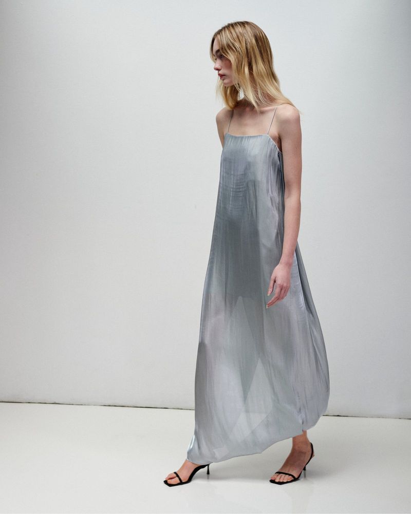 Model wearing a sheer silver spaghetti-strap maxi slip dress and black kitten heels against a white studio sweep.