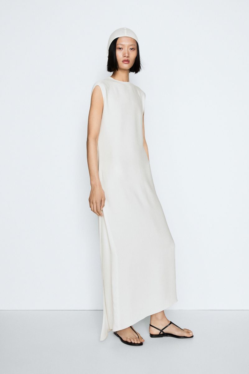 Model wearing a long white sleeveless dress and cap with black sandals standing against a white seamless background